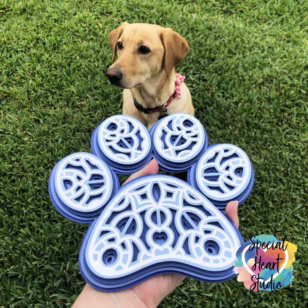 Layered cardstock paw print mandala in blue and white with yellow lab in background in grass