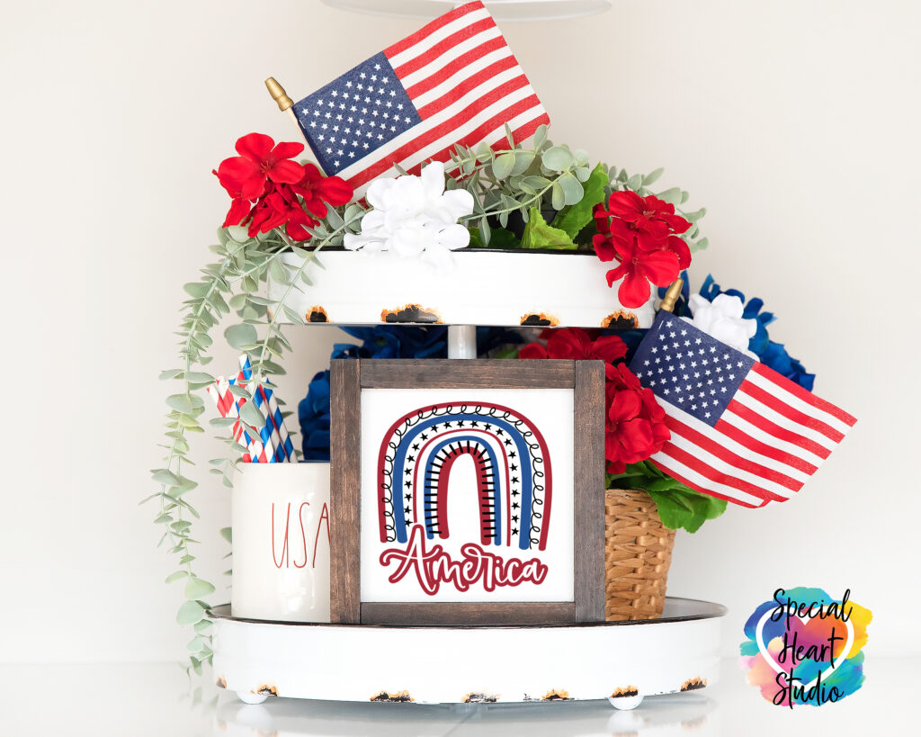 Two tier tray with wood sign in the middle. Sign has boho rainbow in red white and blue with the word America below.  Also on the tray are flowers, greenery and two small US flags.