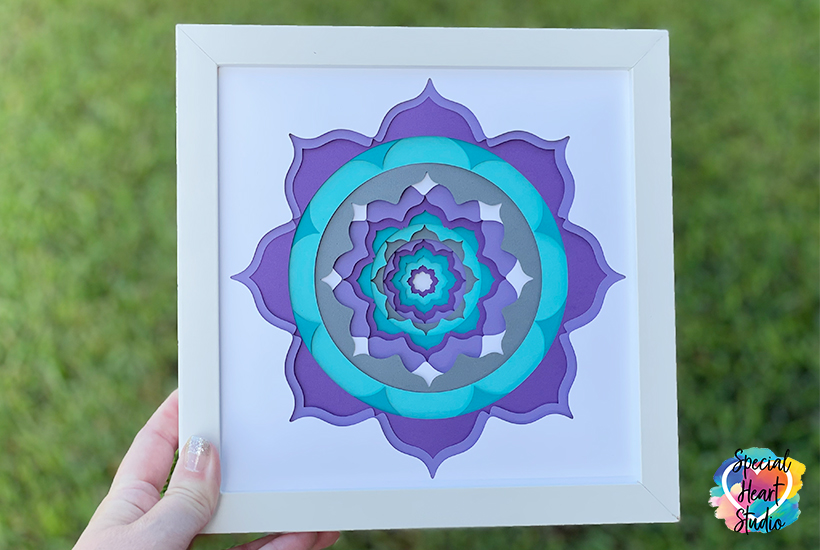 Square white framed shadowbox with layered mandala cut from white, purple and greens.