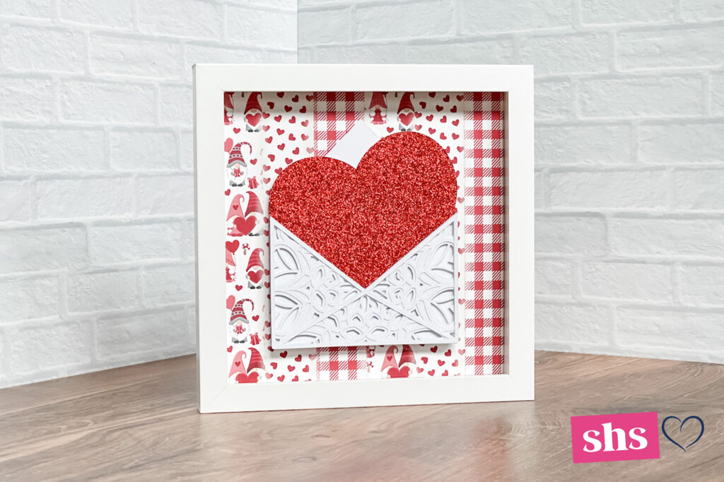 shadowbox frame with Valentine print background. In front is a layered mandala white enevlope with a red glittter heart coming out.