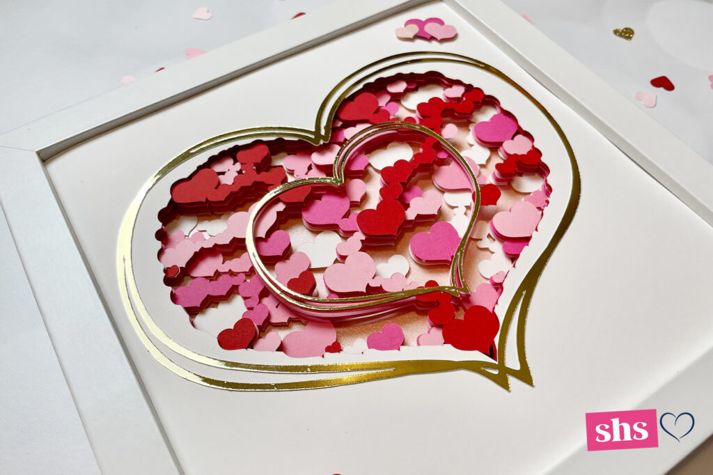 Square shadowbox in white frame with hearts cut from red and various shades of pink hearts.