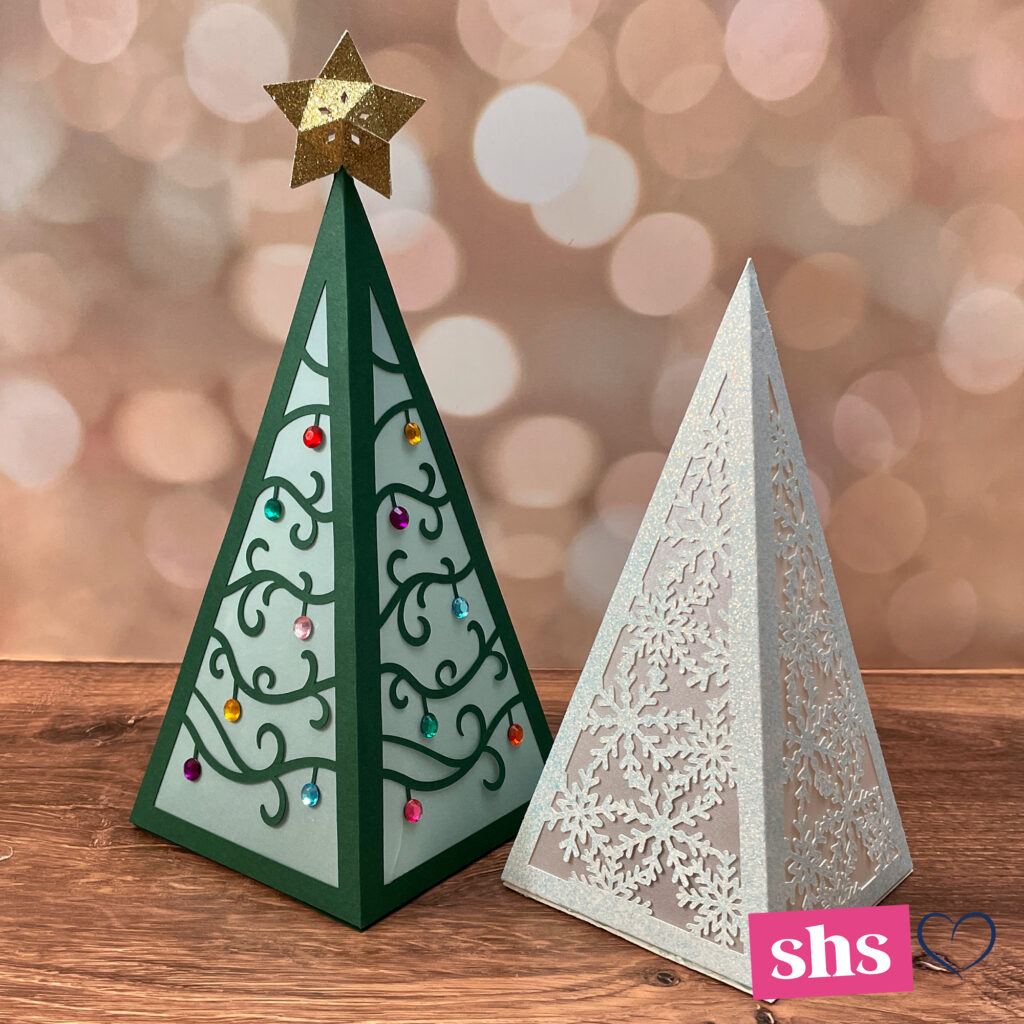 Green paper pyramid tree with gold glitter star on top next to a white glitter pyramid with snowflakes.  Both are luminaries.